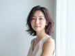 © Ganesha - Close-up portrait of a beautiful, smiling middle-aged Japanese woman standing by the window, touching her neck with one hand and looking at the camera. She is wearing a light grey tank top. generated