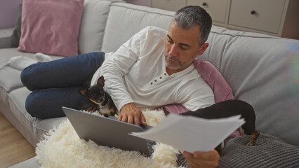 A middle-aged hispanic man working on a laptop in a living room, with two chihuahuas by his side, creating a cozy and productive home environment.