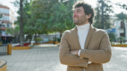 Wall Mural - Handsome hispanic man with a beard, smiling in an outdoor park setting, standing with arms crossed.