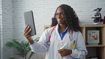 Wall Mural - A young african american woman with curly hair, smiling in a clinic while using a tablet during her work as a doctor.