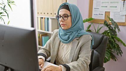 Sticker - A professional muslim woman wearing a hijab works attentively at her computer in a modern office setting.