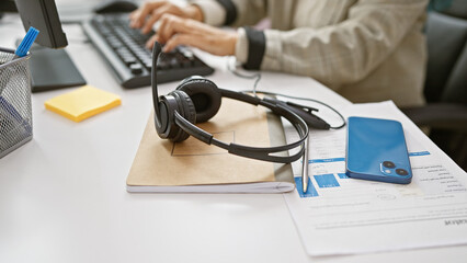 Focused, mature woman typing on a computer in a modern office with headphones and a smartphone on her desk.