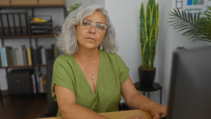 Canvas Print - Woman with grey hair working in an office, looking at a computer screen surrounded by plants and folders