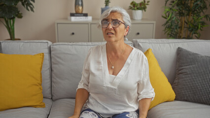 Poster - Grey-haired woman sitting on a sofa in a living room with yellow pillows, wearing a white blouse and glasses, looking concerned