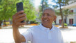 © Krakenimages.com - Grey-haired hispanic man smiling while taking a selfie in an urban park setting on a sunny day outdoors.