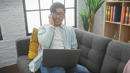 A young asian man multitasks with a phone call and laptop in a modern living room.