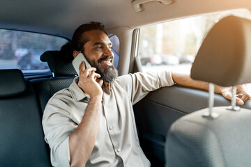 A cheerful man with a beard is engaged in a phone call from the back seat of a car, enjoying the warm sunlight streaming through the window on a bright day.