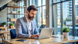 © Tatar - A man working on a laptop in an office with large windows and a plant next to the laptop.