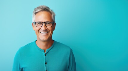 A mature man with gray hair and glasses stands against a vibrant turquoise background, smiling brightly and exuding warmth and wisdom.