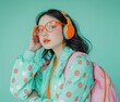 © grigoryepremyan - A young Asian woman with backpack, and orange glasses, listening to music with headphones on an isolated pastel background.