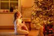 © Irina Schmidt - Cute little girl wearing nightgown and sitting by a beautiful Christmas tree. Christmas and New Year concept. The atmosphere of magic, lights, sparkles around. Happy child with gift looking at tree.
