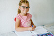 © Irina Schmidt - A young girl with glasses, sitting at a table, is focused on writing in her workbook. Child practicing writing numbers with a pencil, engaged in learning. Elementary school, education.