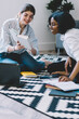 © BullRun - Happy cheerful multicultural female students feeling great from learning togetherness in apartment with stylish interior sitting on floor reading and discussing information from literature book