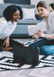 © BullRun - Happy smiling women enjoying teamwork in cozy home interior while sitting on apartment floor with laptop computer and analyzing information from textbook for education during warm conversations