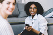 © BullRun - Portrait of cheerful afro american female blogger in optical spectacles looking at camera with cellular phone in hand sitting on apartment floor with millennial caucasian friend on blurred frontage