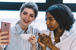 © BullRun - Attractive black skinned girl together with cheerful caucasian friend posing on front camera of smartphone to make cool selfie for publication in social networks, happy women taking photo on cellular