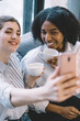 © BullRun - Happy two women friends smiling at front camera of digital smartphone and making photos, cheerful multicultural girls taking selfie on cellphone at home interior during conversations with coffee