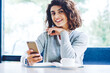 © BullRun - Portrait of positive good looking hipster girl holding mobile phone in hand and smiling at camera during coffee time on publicity area, happy with smartphone gadget enjoying leisure indoors