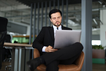 Wall Mural - A man in a suit is sitting in a chair with a laptop in front of him. He is focused on his work, possibly typing or browsing the internet. Concept of professionalism and productivity