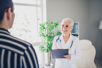 Wall Mural - Portrait of aged woman professional doctor speak patient write clipboard wear white coat workplace office indoors