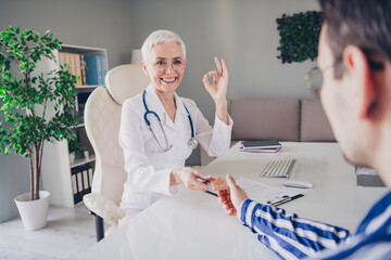 Wall Mural - Portrait of aged woman professional doctor give patient pills show okey symbol wear white coat workplace office indoors