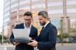 © Panacea Stock  - Two businessman on the street, deeply engrossed, eyes fixed on their laptop screen. Two handsome young businessmen in classic suits using laptop. Business men team using laptop outdoor. Business man