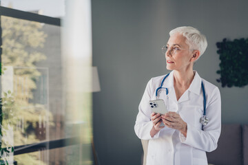 Poster - Portrait of aged woman professional doctor hold smart phone look window think wear white coat workplace office indoors