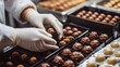 © Luka - A chocolate factory worker's hands in white gloves placing chocolate truffles into black boxes, with chocolate bonbons in the background