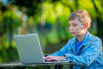 Teen boy with a laptop sits behind a table in a park. School boy is typing on computer outdoors. Young boy studying online with a computer, outdoors. Boy on online lessons while outside.