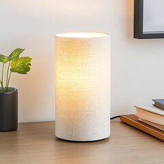 Wall Mural - A white cylindrical lamp with a fabric shade sits on a wooden table next to a plant, books, and a framed picture.