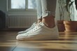 © YURY YUTY - Person wearing white sneakers stands on hardwood floor near potted plants inside a sunlit room.