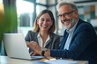 © Vadym - Two middle-aged business professionals smiling confidently at office desk. Man and woman work together, use laptop for project. Happy and successful colleagues collaborate, manage business.