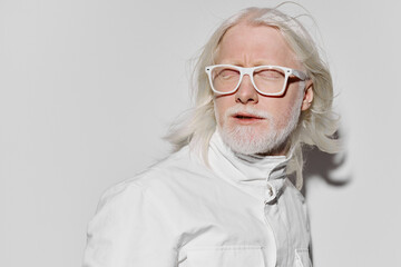 Portrait of an elderly man with white hair and beard wearing white glasses against a plain white background, showcasing a unique fashion style and serene expression