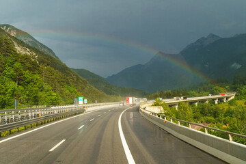 A scenic drive along a winding highway with a vibrant rainbow arching over lush green mountains after a refreshing rain