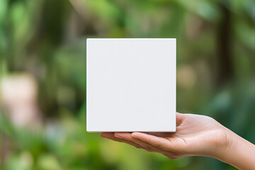 A delicate hand is offering blank white cube for creative purposes, set against lush green background that evokes sense of tranquility and inspiration