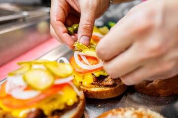 Wall Mural - Close-up of hands placing pickles on a freshly made burger with lettuce, tomato, and cheese. Perfect for food preparation, culinary arts, and fast food themes. High-quality, appetizing, and vibrant.