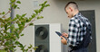 © StockMediaSeller - A technician uses a tablet to adjust settings on an external heat pump unit outside a home.