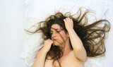 sexy nude woman with disheveled hair lies on a white pillow, her expression distant and reflective. Her hand lightly touches her face, suggesting vulnerability, emotion, or contemplation