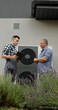 © StockMediaSeller - A technician discusses heat pump functionality with a homeowner in front of the unit, using a tablet for reference.