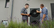 © StockMediaSeller - A technician and homeowner discuss heat pump maintenance, referencing a tablet near the unit.