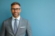 © pham - A professional man with a beard and glasses poses confidently in a gray suit against a blue backdrop, showcasing a friendly demeanor