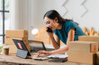 © Kritdanai - E-commerce Hustle: Young Asian entrepreneur managing orders and logistics on phone and tablet, surrounded by shipping boxes in her home office.