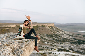 Wall Mural - Serene woman in hat sitting on mountain peak, contemplating the vast expanse of nature