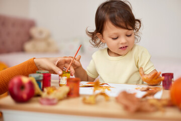 smiling little child girl painting her hands and autumn leaves. baby girl drawing hands in yellow pa