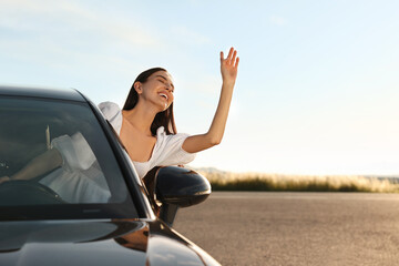 Wall Mural - Smiling young woman leaning out of car window outdoors. Enjoying trip