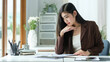 © wattana - Young woman with long dark hair is seated at a bright, modern desk, thoughtfully reviewing documents.