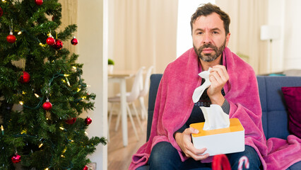 Man feeling unwell and blowing his nose while seated on a couch during the holiday season