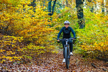 Wall Mural - Middle aged woman wearing black sporty jacket, black cycling pants and white bike helmet riding bicycle in forest in autumn scenery. Front view.	