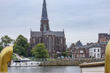 St. Martin Church on banks of Meuse river against grey sky, yacht anchored at small pier on promenade, cityscape, buildings in background, cloudy day in Maastricht, South Limburg, Netherlands