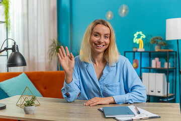 Young businesswoman smiling friendly at camera and waving hands gesturing hello, hi, greeting or goodbye, welcoming with hospitable expression at home office workplace desk. Happy girl in living room.